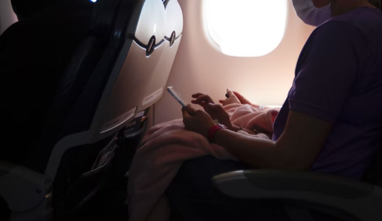 Person using a smartphone while seated on an airplane next to a window, wearing a mask and holding a blanket.