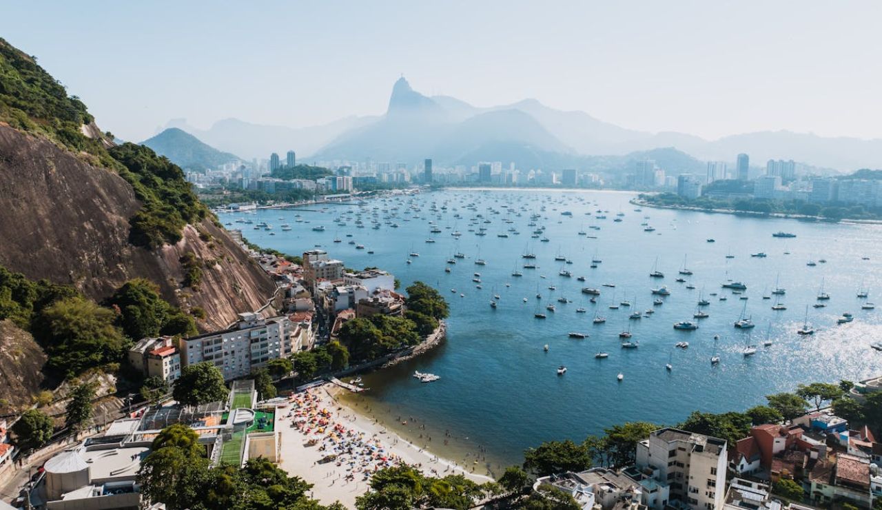 Aerial view of Rio de Janeiro's coastline, with boats on the water, a beach filled with umbrellas, and mountains in the background.