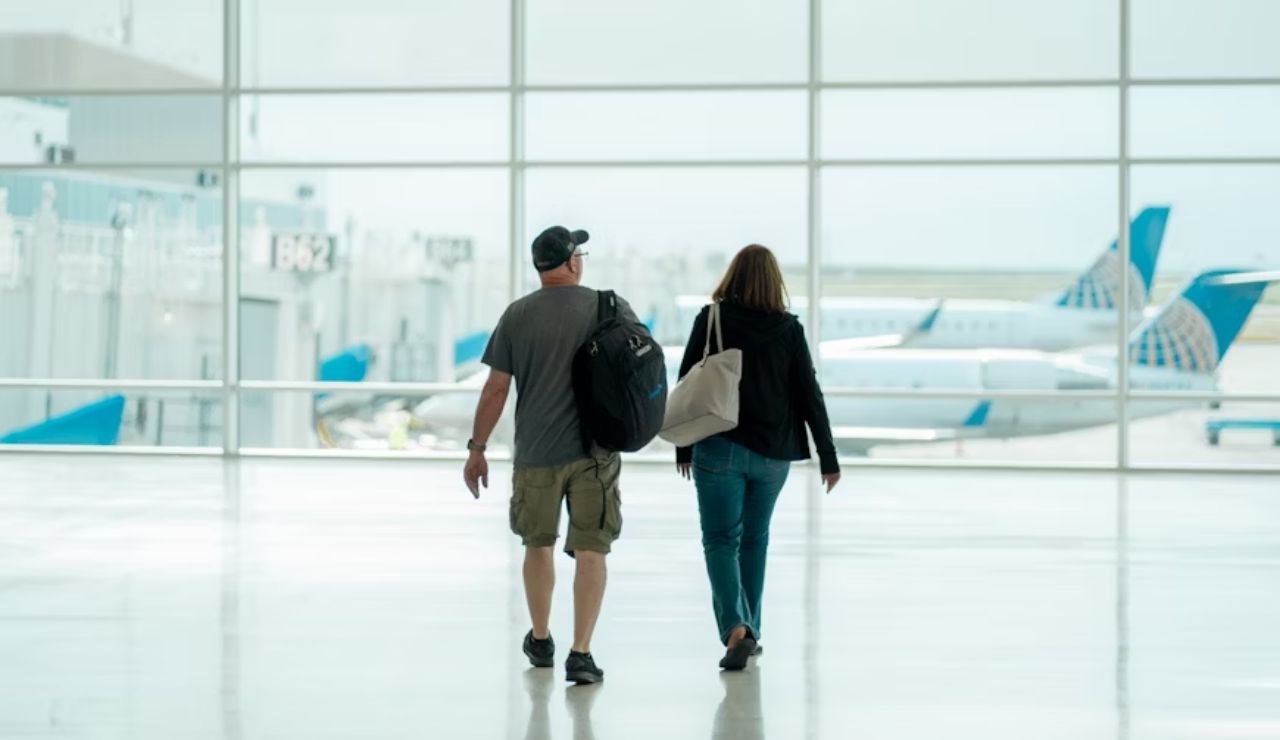 Two travelers walking through a bright airport terminal with planes visible outside the large glass windows.