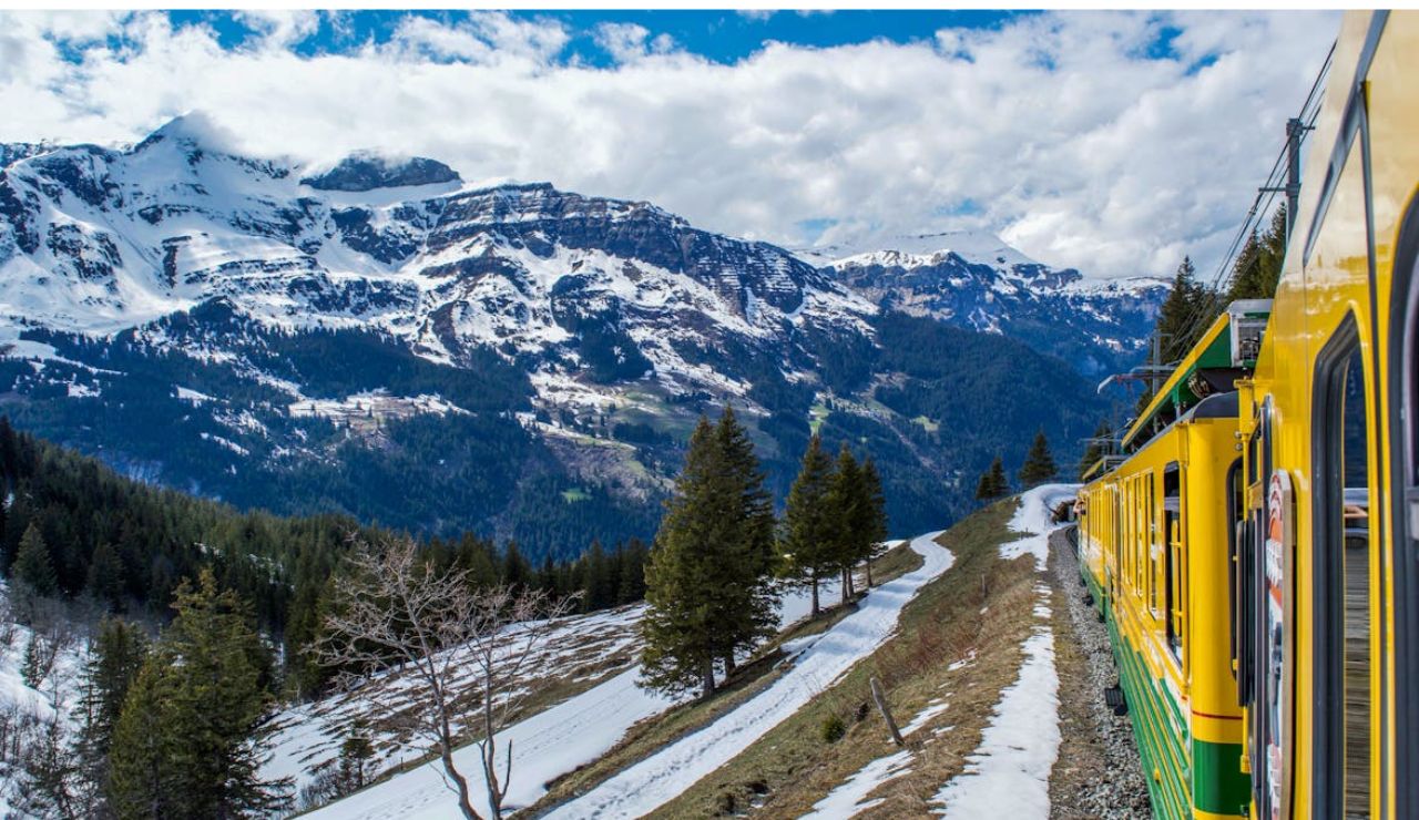 Yellow train traveling through snowy alpine mountains with scenic forest views.