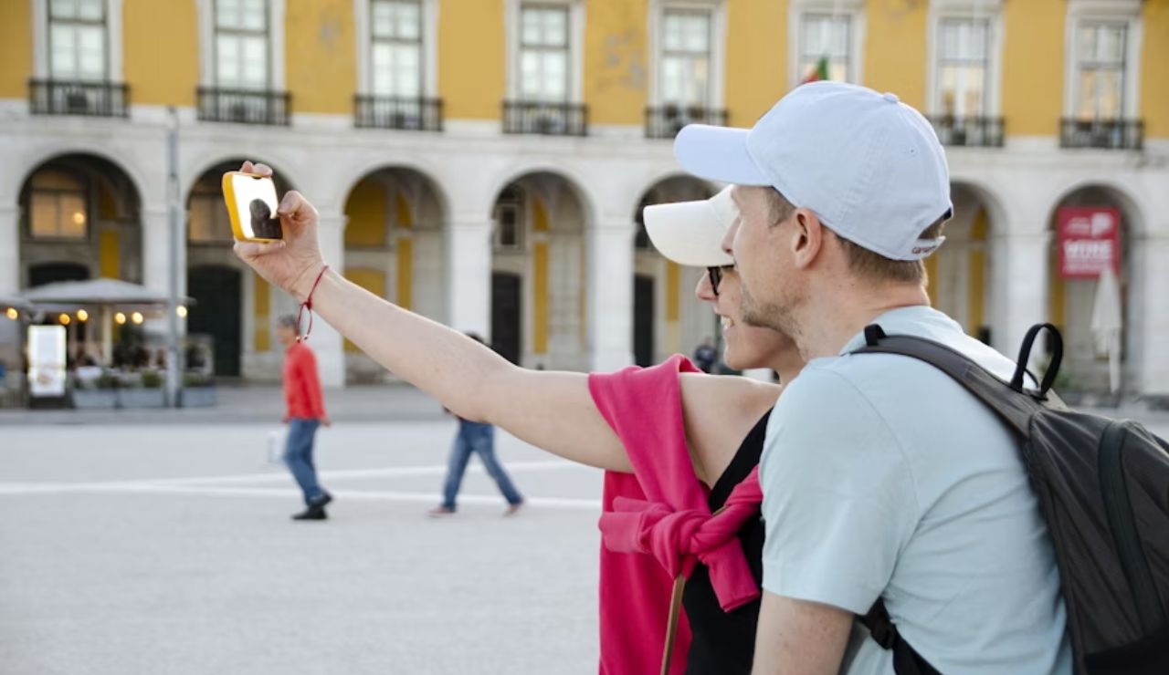 A couple taking a selfie together in a lively outdoor setting, with a yellow building in the background.