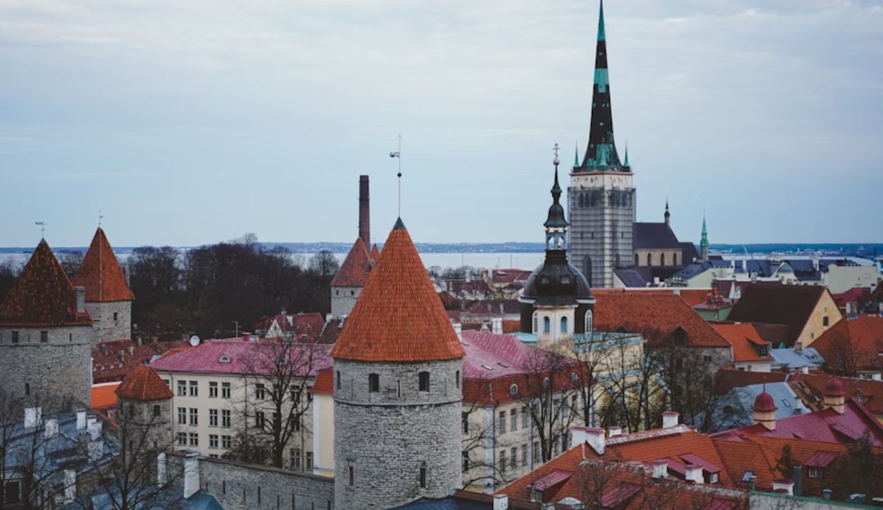 View of a historic European city with red-roofed buildings, stone towers, and a tall church spire near the coastline.
