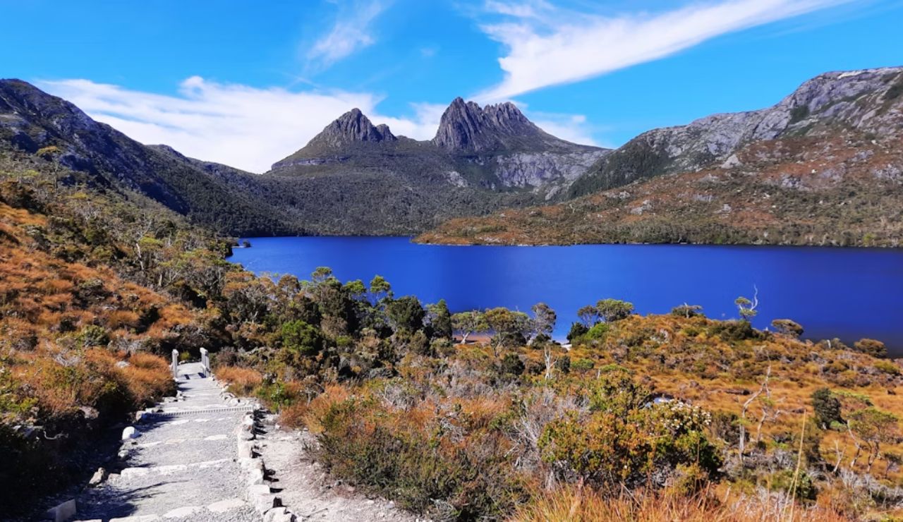 Scenic hiking trail leading to a deep blue mountain lake, surrounded by rugged peaks and alpine vegetation under a vibrant blue sky.