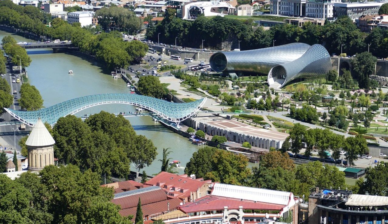Aerial view of a modern glass pedestrian bridge and tubular buildings spanning a river in a green urban park setting.