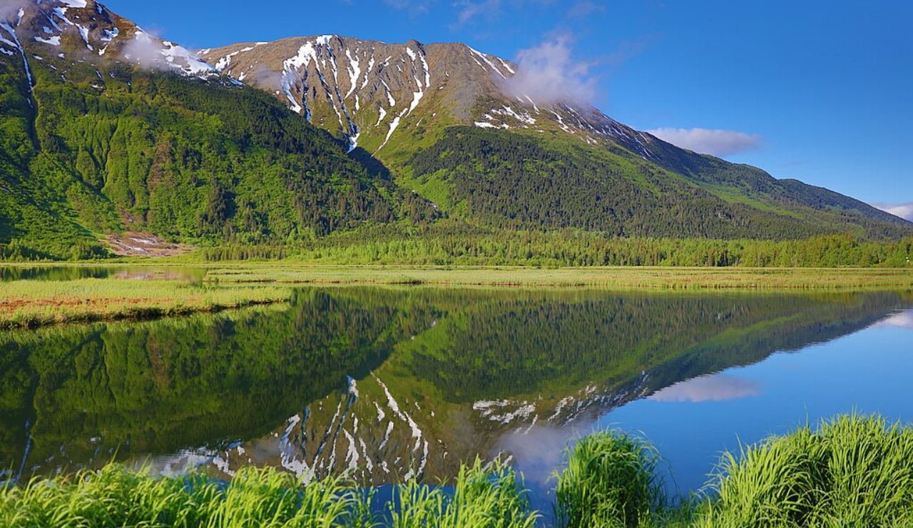 A calm lake reflects snow-capped mountains and lush green forest at Eagle River in Alaska under a clear blue sky.
