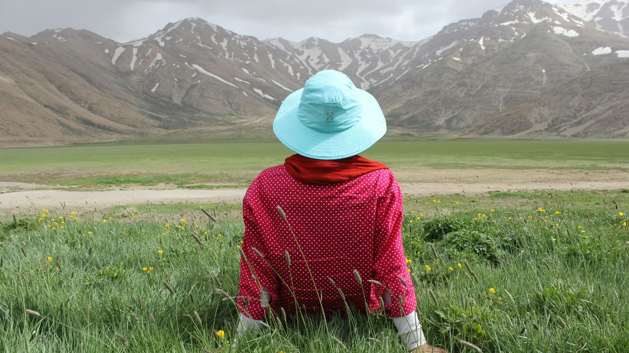 Person wearing a turquoise sunhat and red shirt sitting on green grass, facing snow-capped mountains under a cloudy sky.