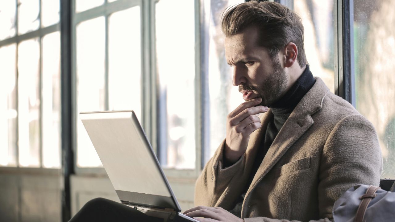 Man in a blazer working thoughtfully on a laptop by a window.