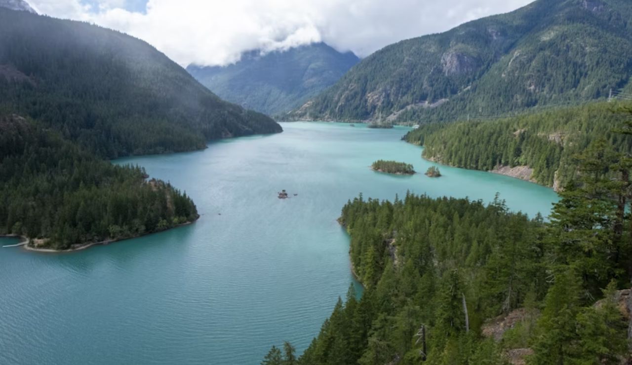 Scenic view of a turquoise lake surrounded by dense evergreen forests and mountains under a partly cloudy sky.