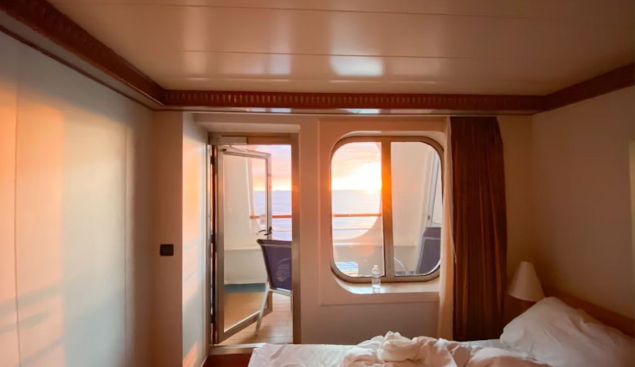 Cozy cruise ship room with an unmade bed, a window, and an open door leading to a private balcony overlooking the ocean at sunset.