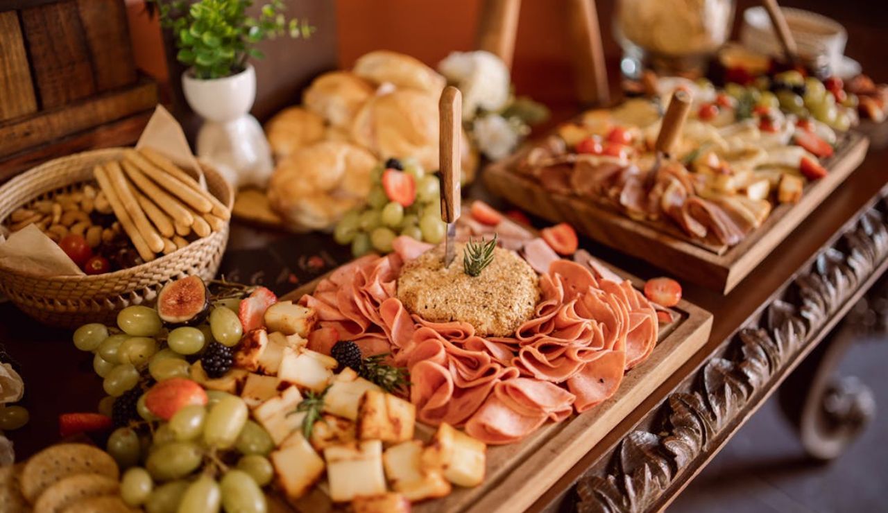 A beautifully arranged charcuterie board with sliced meats, cheese cubes, grapes, figs, and crackers, surrounded by bread rolls and breadsticks.