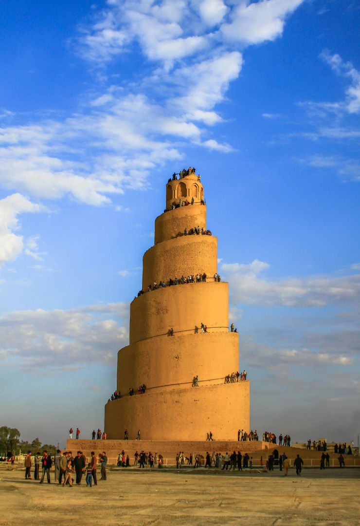 Spiral minaret of the Great Mosque of Samarra in Iraq, with visitors walking along its winding ramp under a bright blue sky.