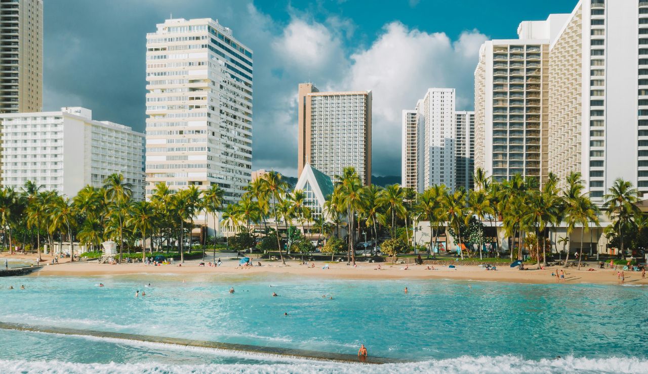 High-rise hotels and palm trees border a sandy beach where people relax by turquoise water under a partly cloudy sky.