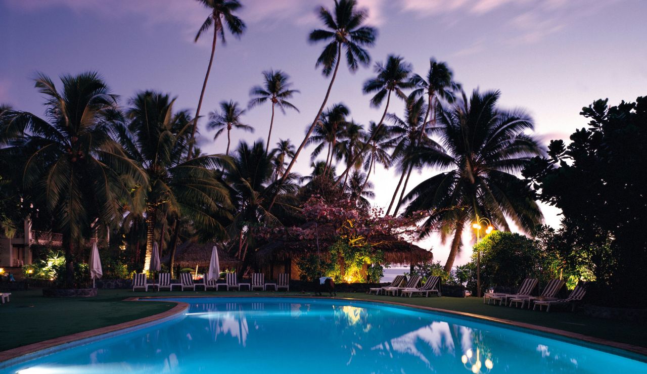 At dusk, lights reflect on a swimming pool surrounded by lounge chairs and tall palm trees beneath a purple sky.