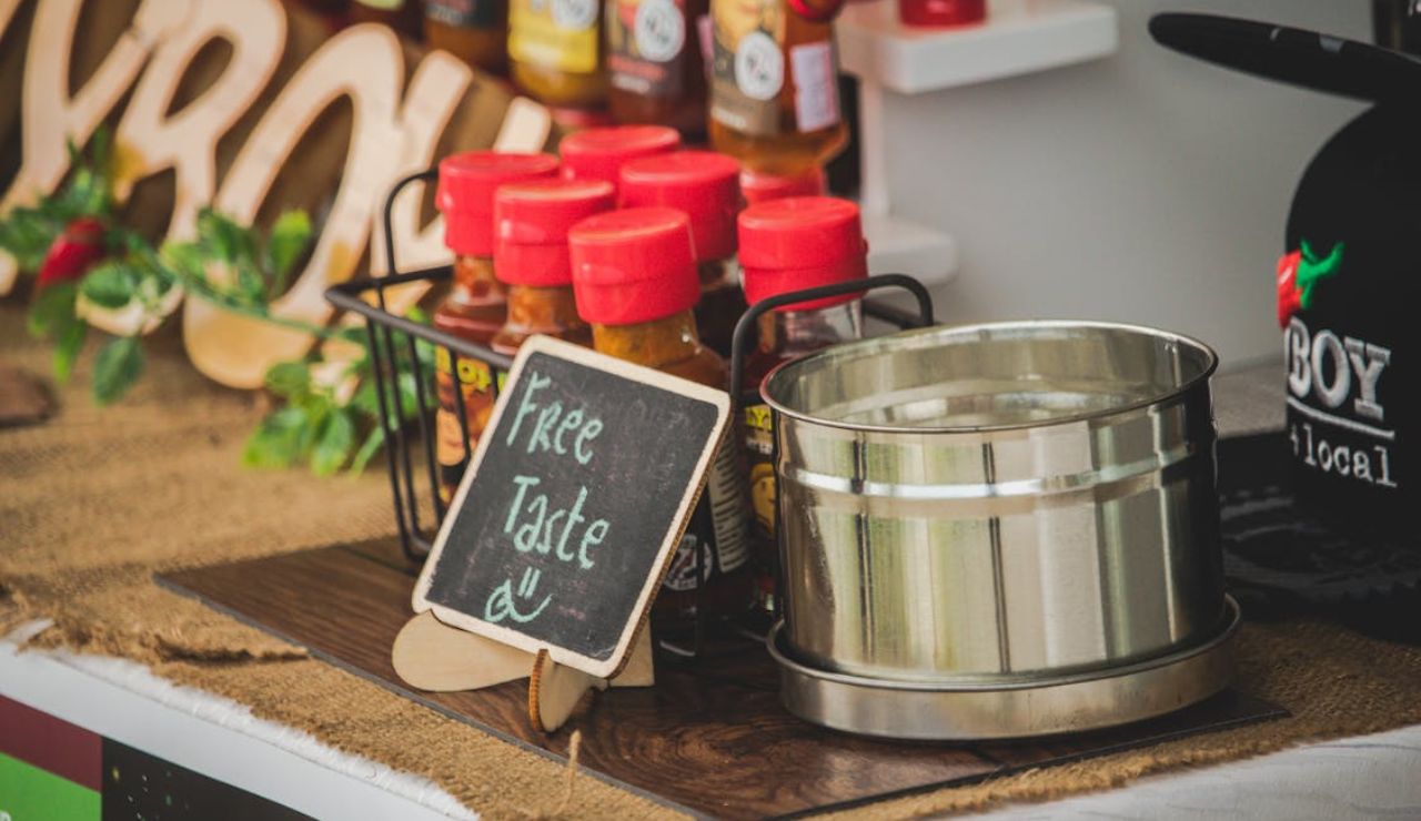 Display of bottled sauces with red caps beside a metal container and a small chalkboard sign that reads "Free Taste :)"