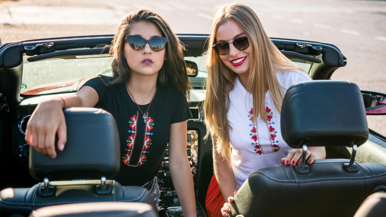 Two women in embroidered shirts and sunglasses sit in a convertible, one dark-haired with her arm on the seat and the other blonde and smiling.