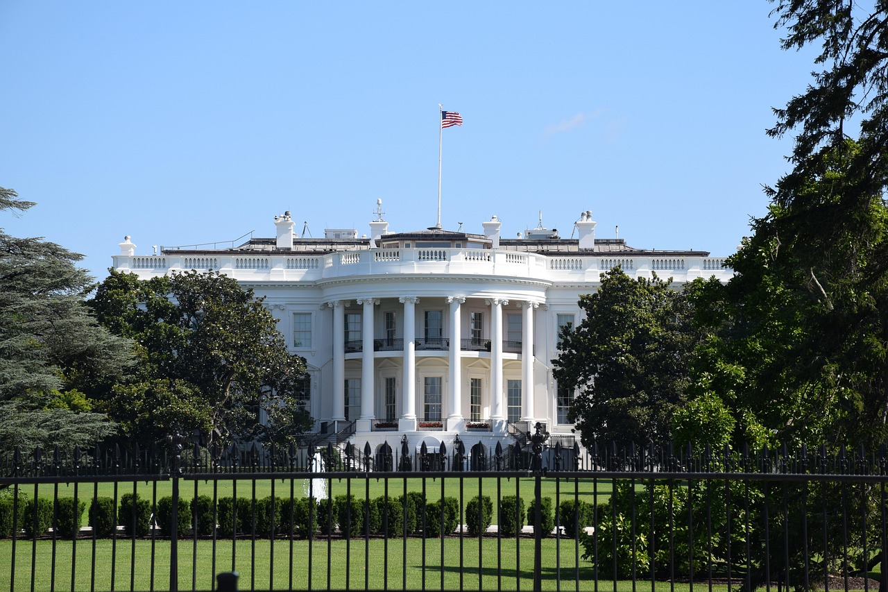 Front view of the White House with the American flag flying above, framed by trees and a black iron fence under a clear blue sky.