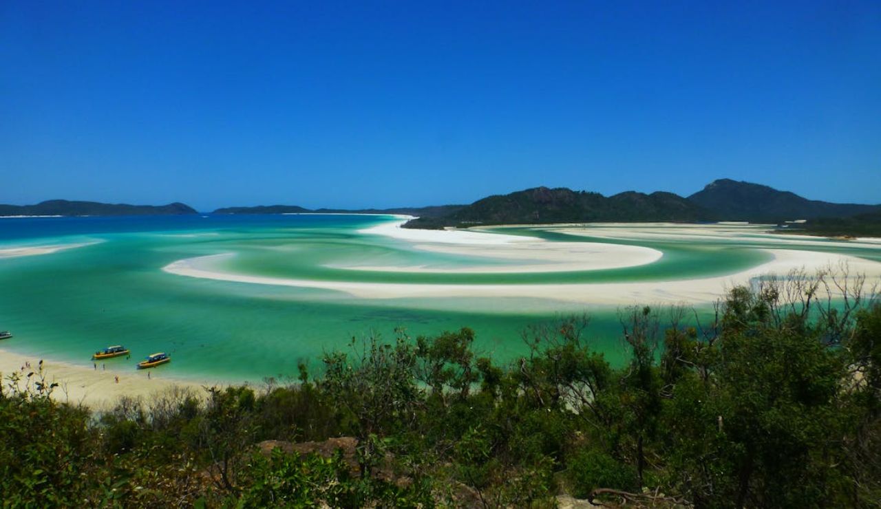 Stunning view of swirling turquoise waters and white sandbars at a tropical beach, with small boats anchored near the shore and forested hills in the background.