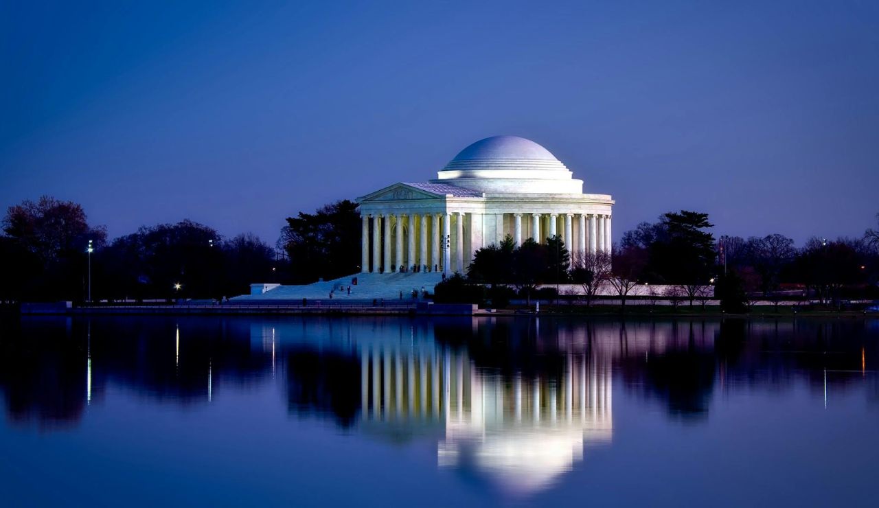 Illuminated Jefferson Memorial at dusk, reflecting in the still waters of the Tidal Basin under a deep blue evening sky.