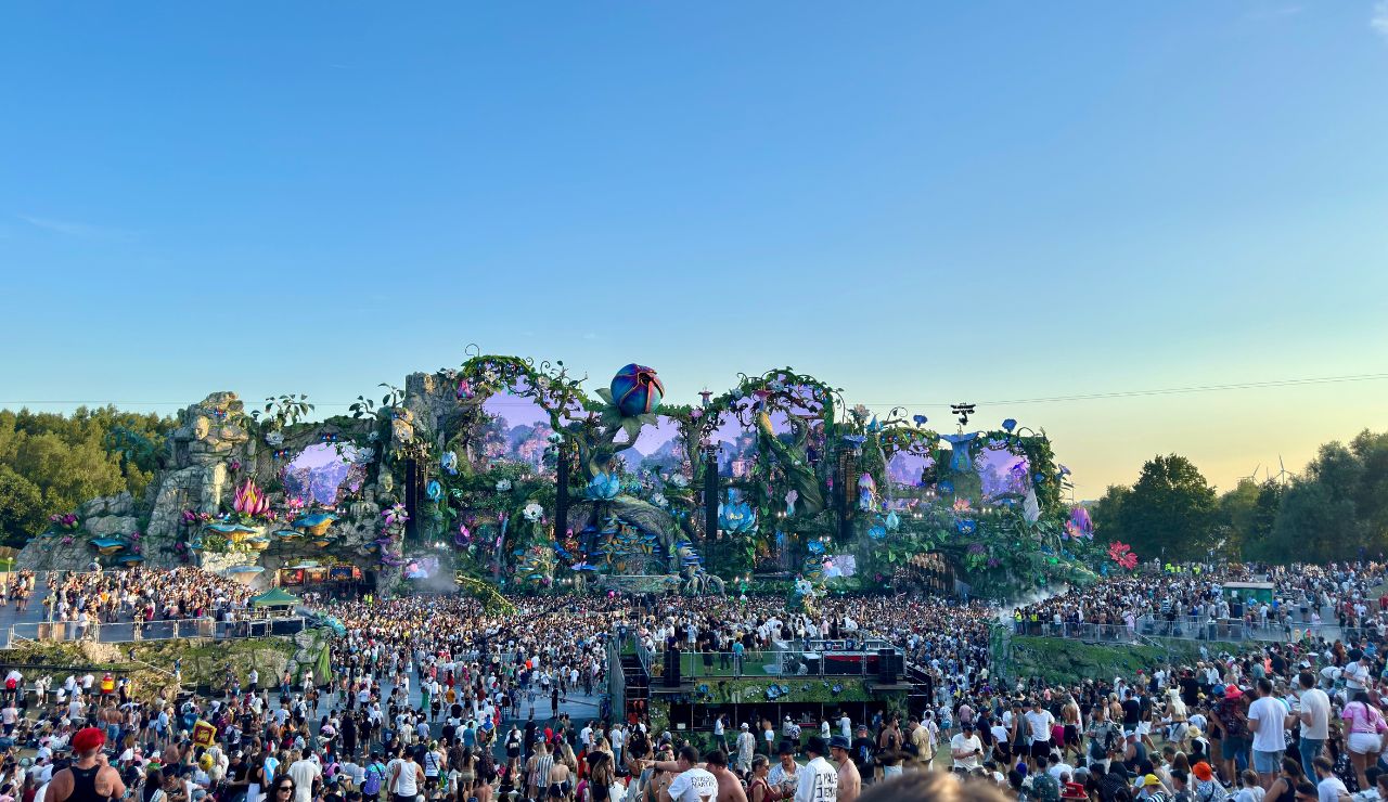 A massive crowd enjoys an outdoor electronic music festival in front of an elaborate, fantasy-themed stage adorned with lights, greenery, and giant props under a clear blue sky.