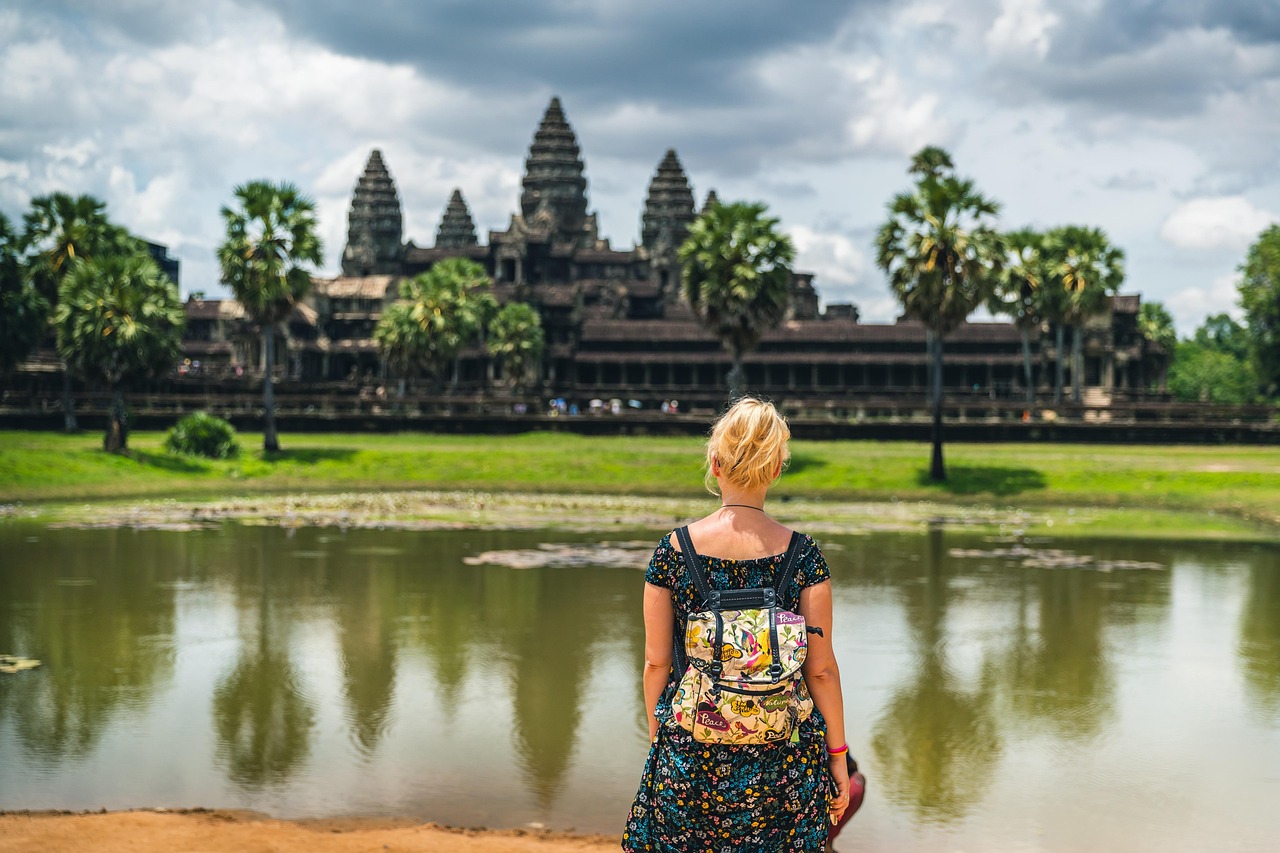 A person with a colorful backpack stands by a pond, facing Angkor Wat beneath a cloudy sky.