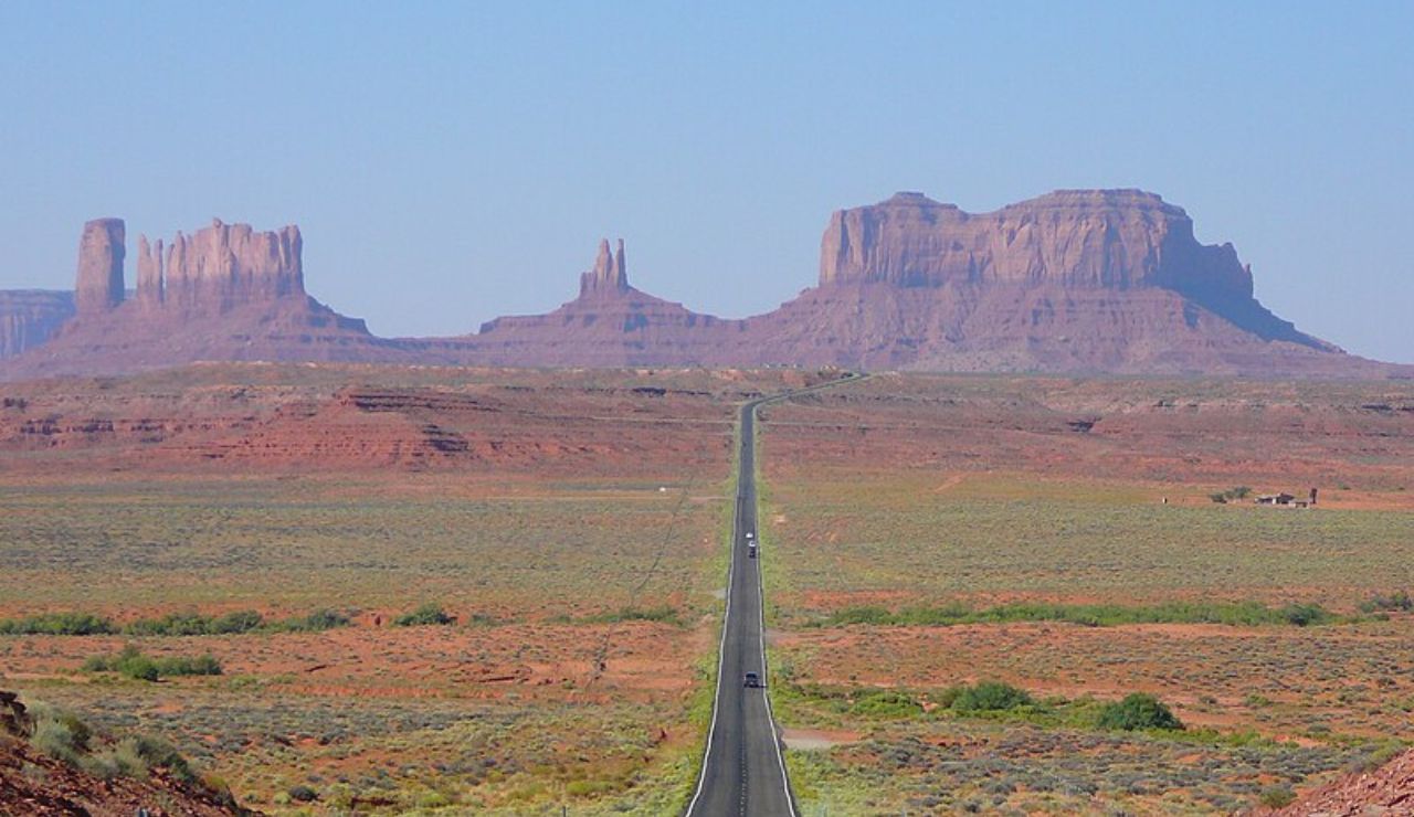 Straight desert highway stretching toward towering red rock formations in Monument Valley under a clear blue sky.