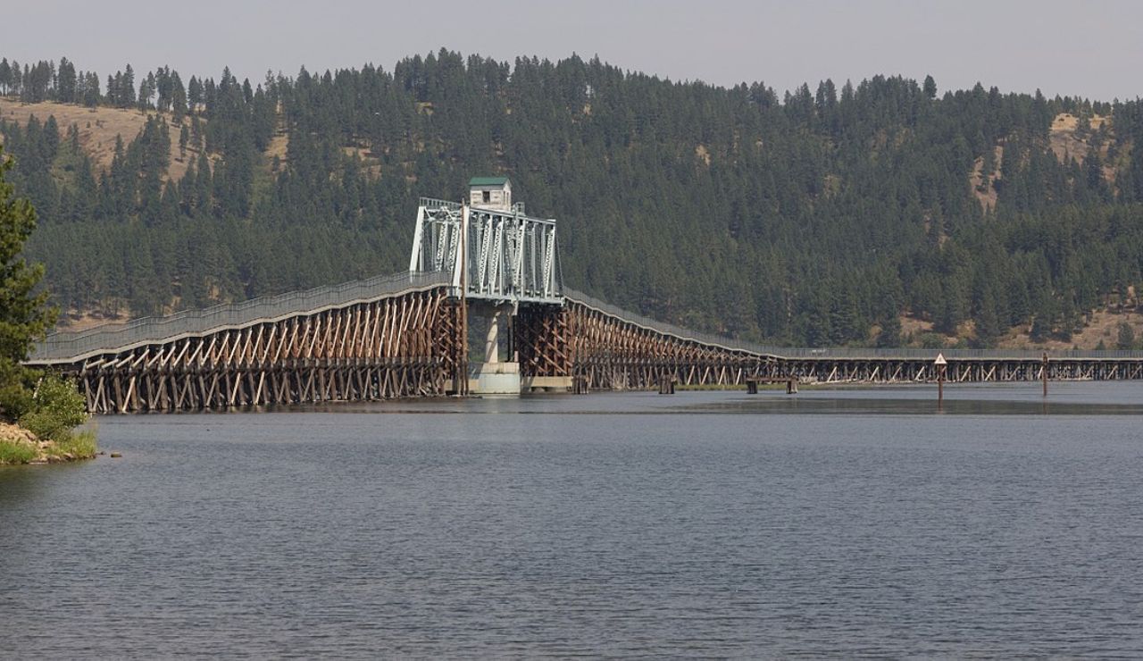Long wooden trestle bridge with a central metal drawbridge spanning a wide river, surrounded by forested hills.