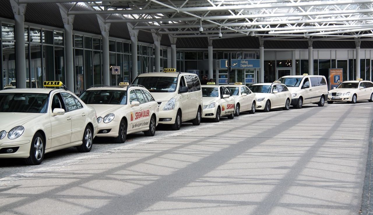 A row of white taxis lined up outside an airport terminal under a covered walkway.