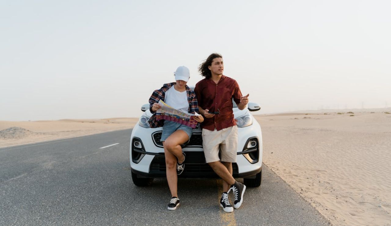 Two travelers stand in front of a white car on a deserted road through the desert, looking at a map and phone while deciding which way to go.