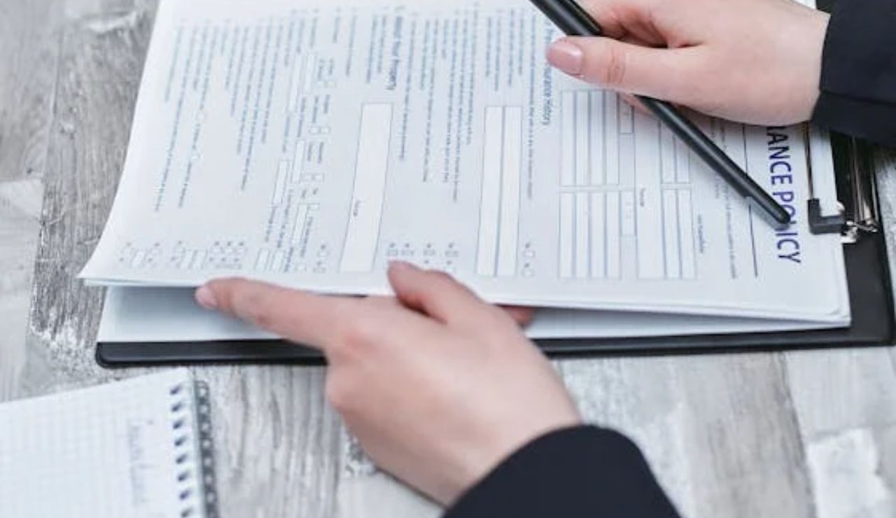 A person filling out an insurance policy form on a clipboard with a pen, seated at a wooden table.