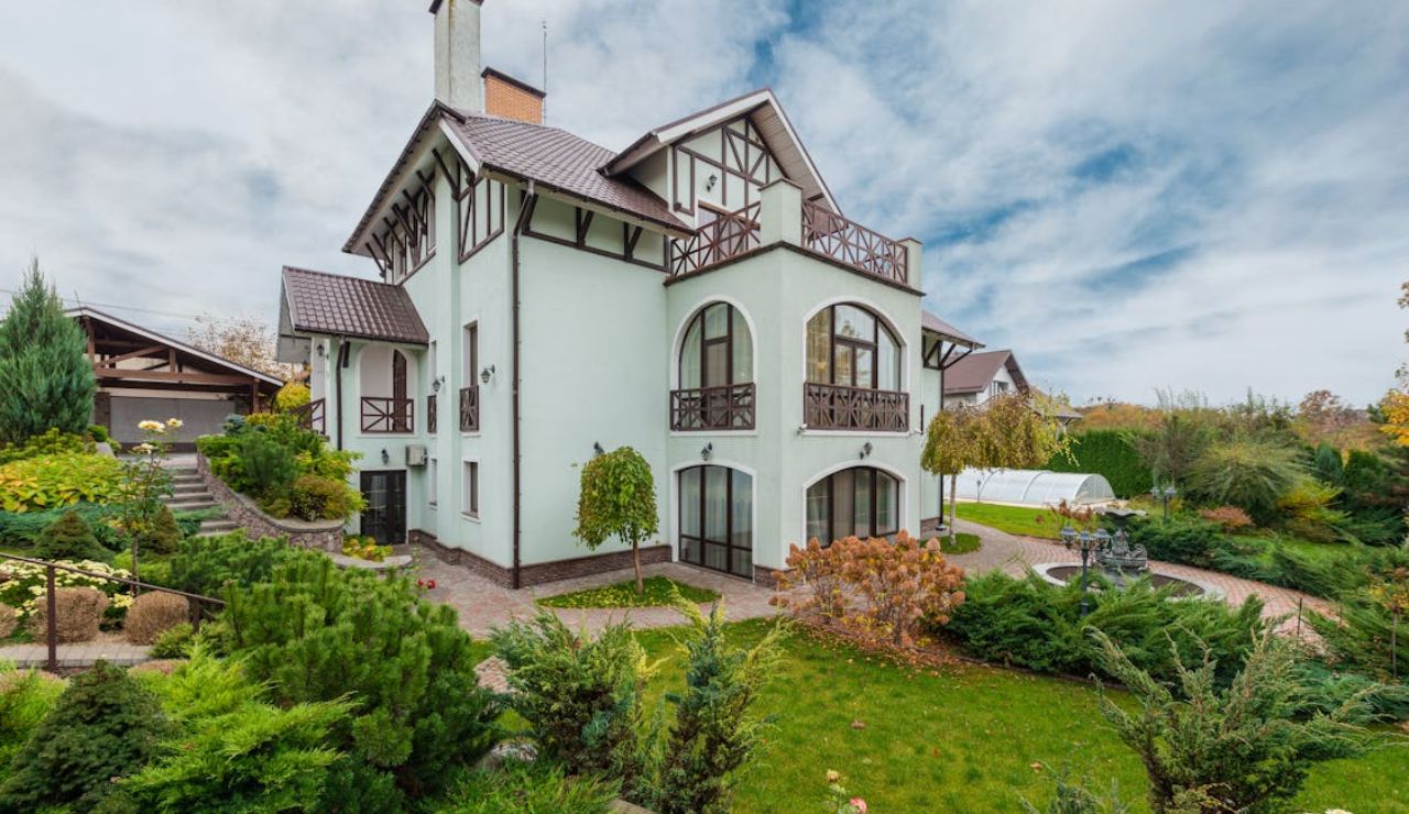 Elegant multi-story house with Tudor-style architecture, surrounded by a landscaped garden featuring shrubs, trees, and a paved walkway.