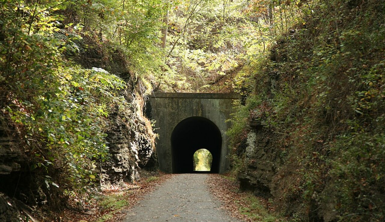 Tree-lined trail leading to a dark tunnel entrance carved into a rocky hillside along a peaceful forest path.