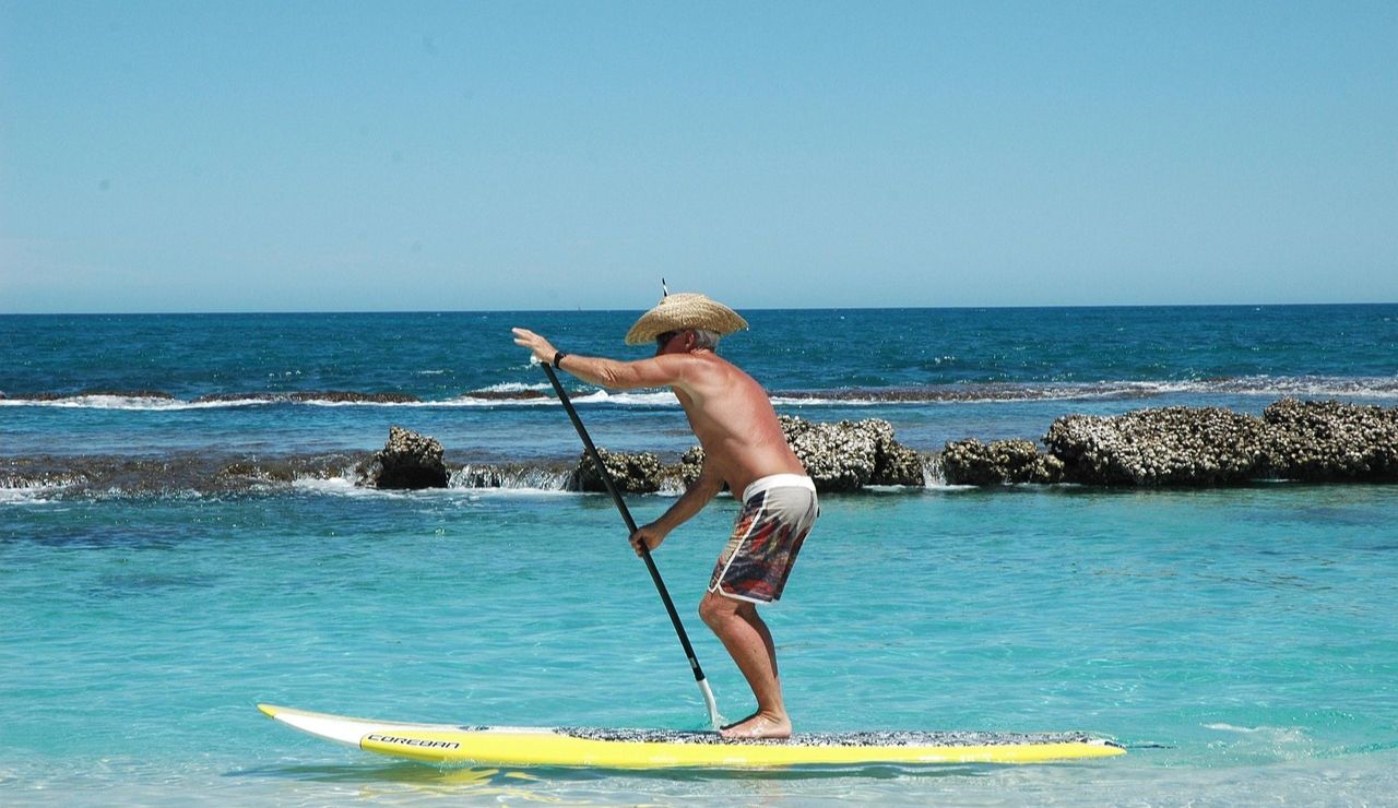 A man in a straw hat paddleboards through crystal-clear turquoise waters near a rocky shoreline.