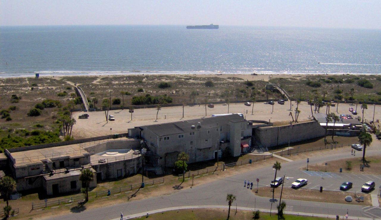 Aerial view of a coastal fort near a beach with a parking lot, walkways to the shoreline, and a large ship in the distant ocean.