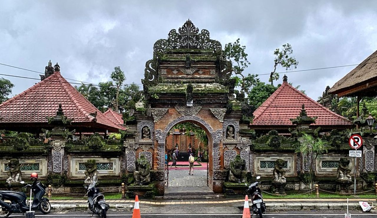 Traditional Balinese temple gate with ornate carvings, flanked by statues and red-roofed pavilions, on a cloudy day in Ubud, Bali.