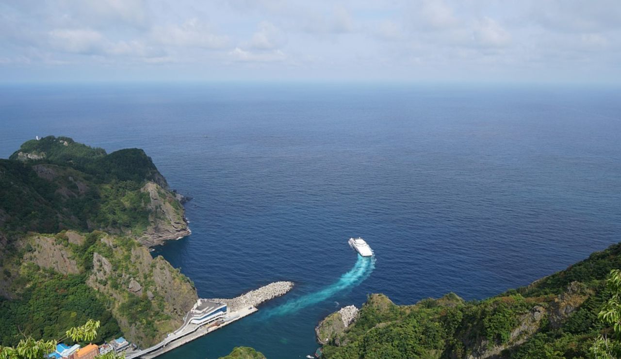 Ferry boat leaving a small harbor surrounded by lush green cliffs, heading into the open ocean under a partly cloudy sky.