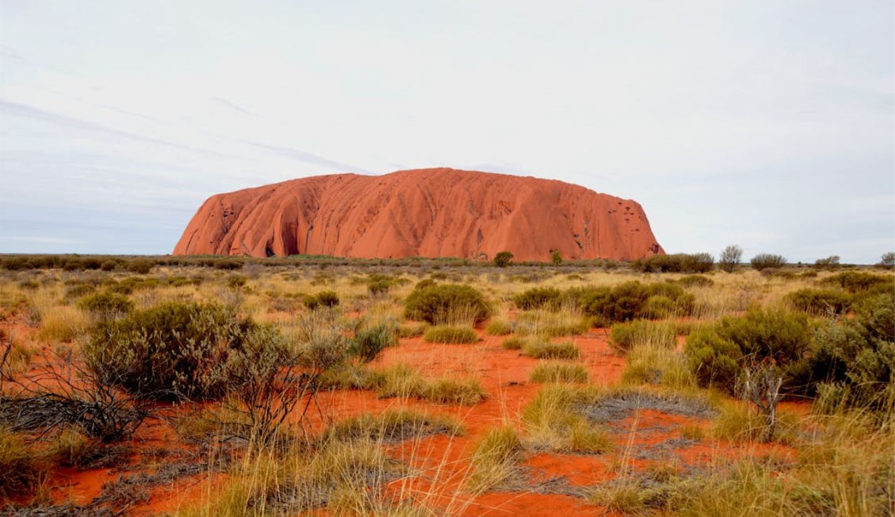 Iconic red sandstone monolith Uluru rising from the arid desert landscape, surrounded by dry grass and scattered shrubs.