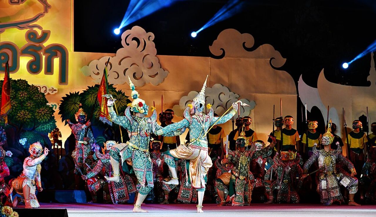 Traditional Thai dance performance on stage featuring elaborately costumed dancers in ornate masks, with dramatic lighting and a painted backdrop.