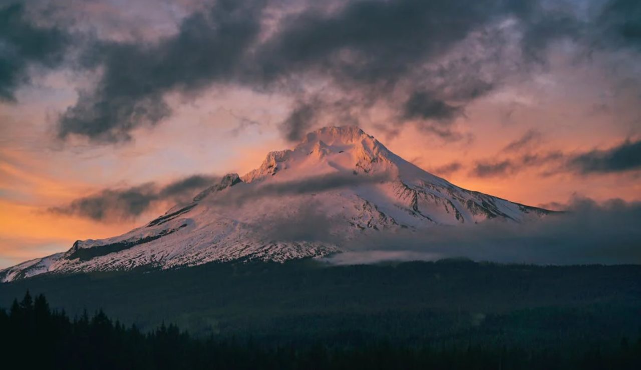 Snow-covered mountain peak at sunset, with dramatic clouds and forest in the foreground.