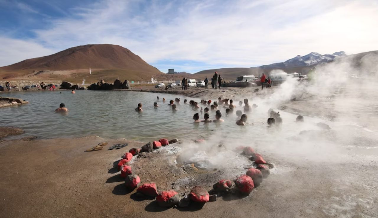 People bathing in a natural hot spring surrounded by mountains, with steam rising from the water under a partly cloudy sky.