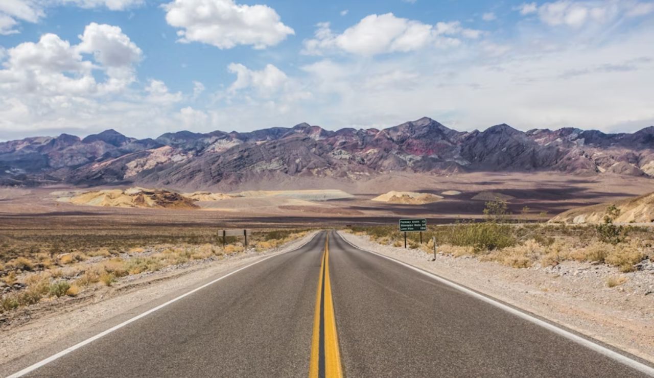 Open desert highway leading toward colorful, rugged mountains under a bright blue sky with scattered clouds.