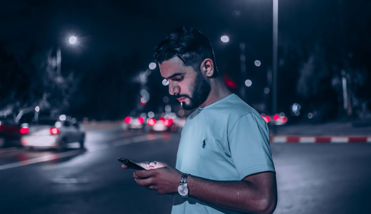 Man standing on a city street at night, looking at his phone with blurred car lights in the background.