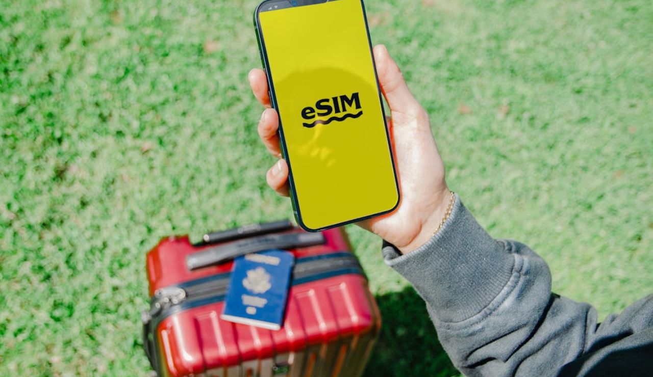 Person holding a smartphone displaying an "eSIM" screen, with a suitcase and passport on the grass in the background.