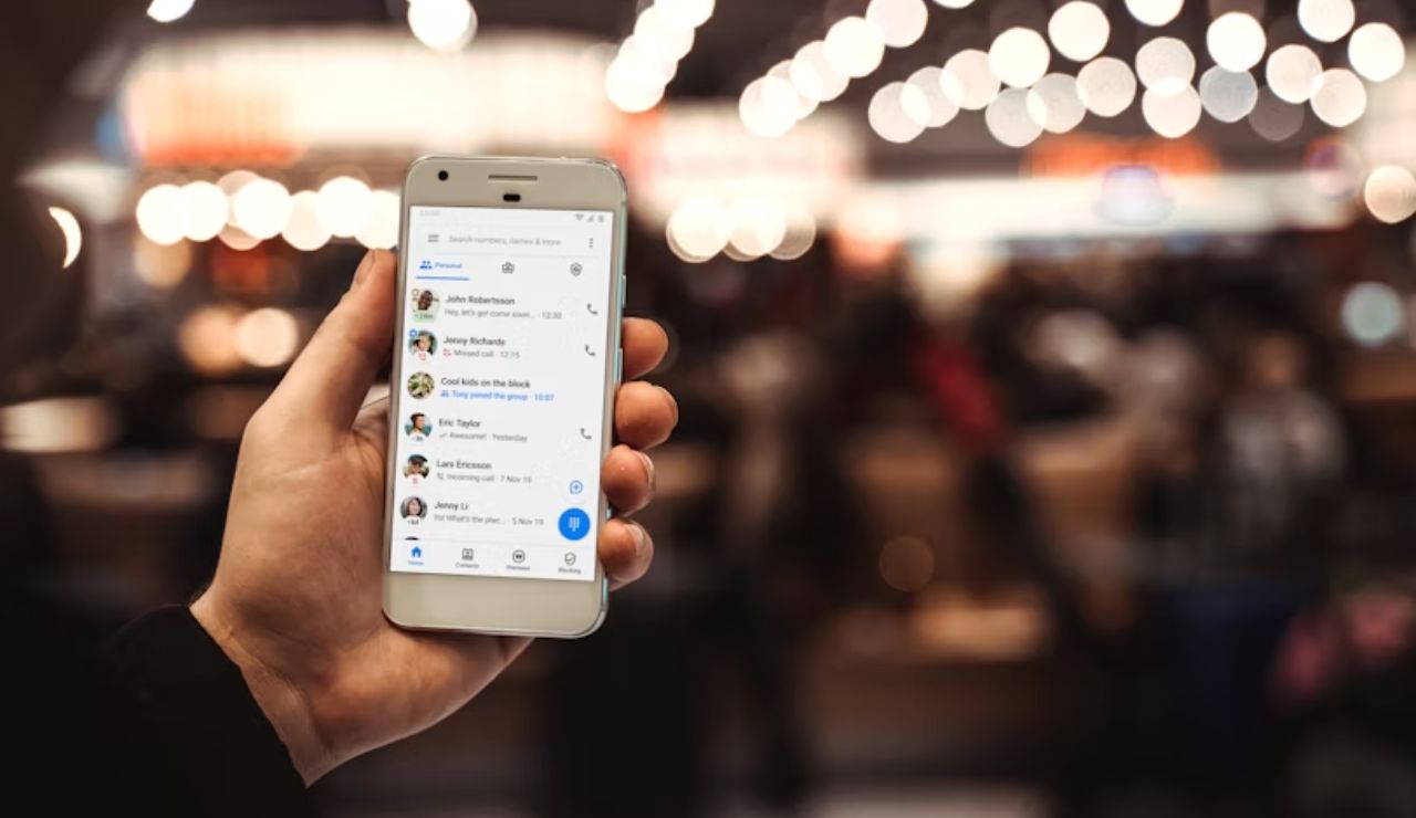 Hand holding a smartphone displaying a messaging app interface, with a blurred crowd and string lights in the background.