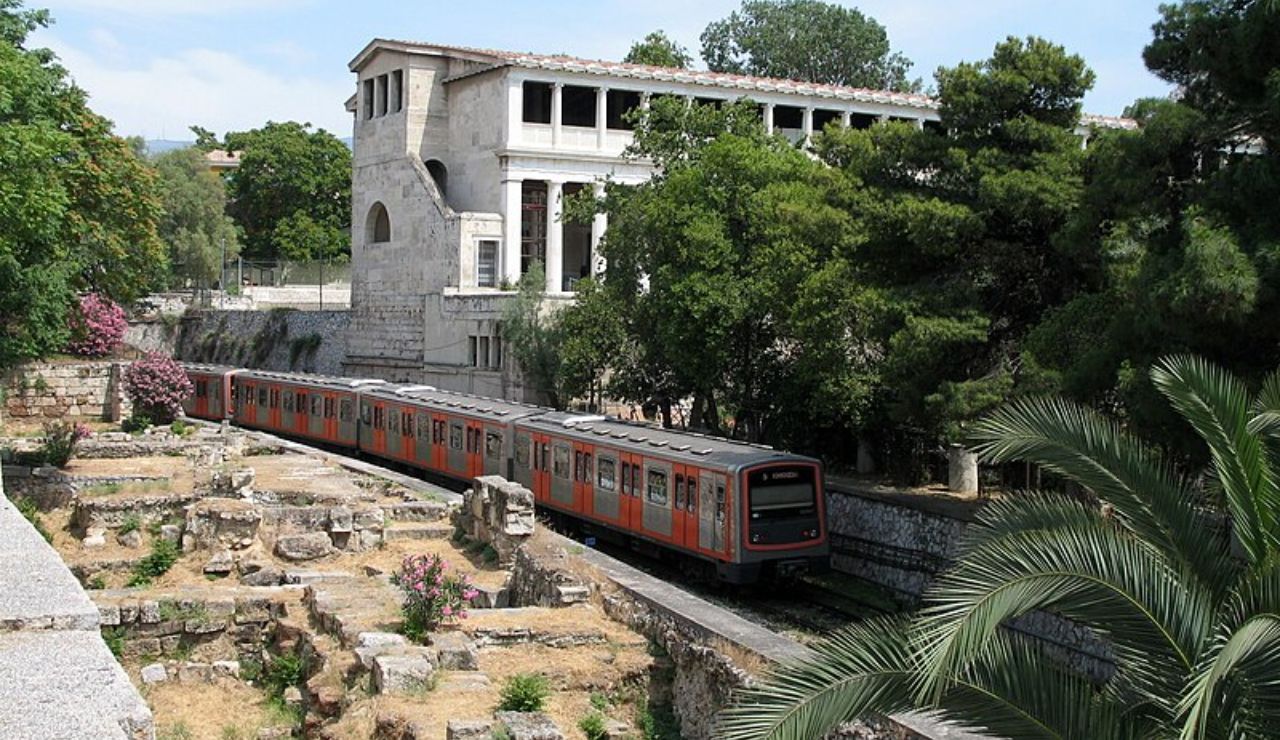 Modern metro train passing through ancient ruins near the Stoa of Attalos in Athens, Greece, surrounded by greenery.