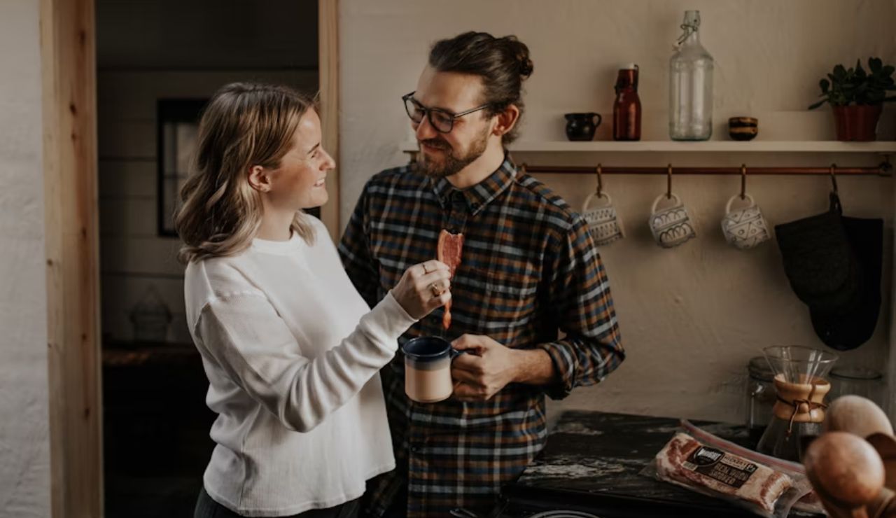 Couple standing in a cozy kitchen, smiling at each other while sharing a piece of food and holding coffee.