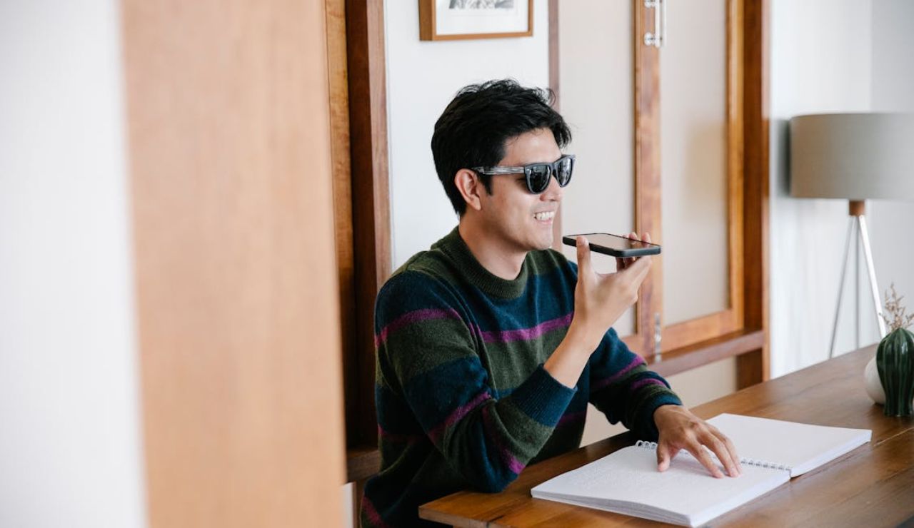 Man wearing sunglasses sitting at a desk, speaking into a smartphone with a notebook open in front of him.
