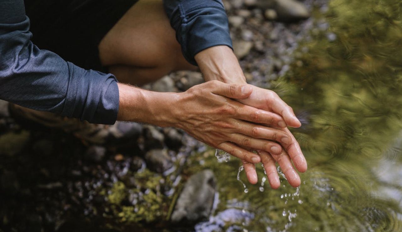 Person rinsing hands in a natural stream, highlighting the importance of not using soap or detergents in water sources.
