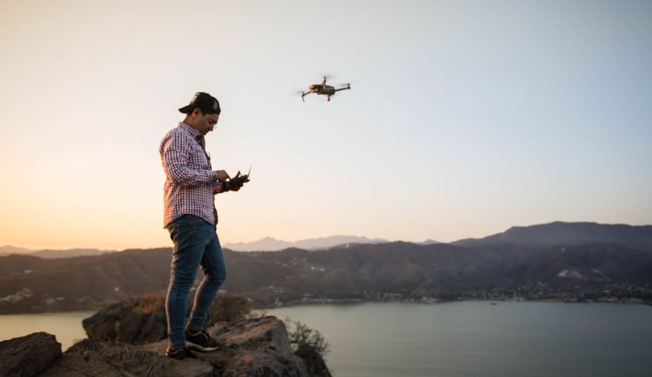 Man standing on a cliff at sunset operating a flying drone with a remote controller, overlooking a lake and mountains.