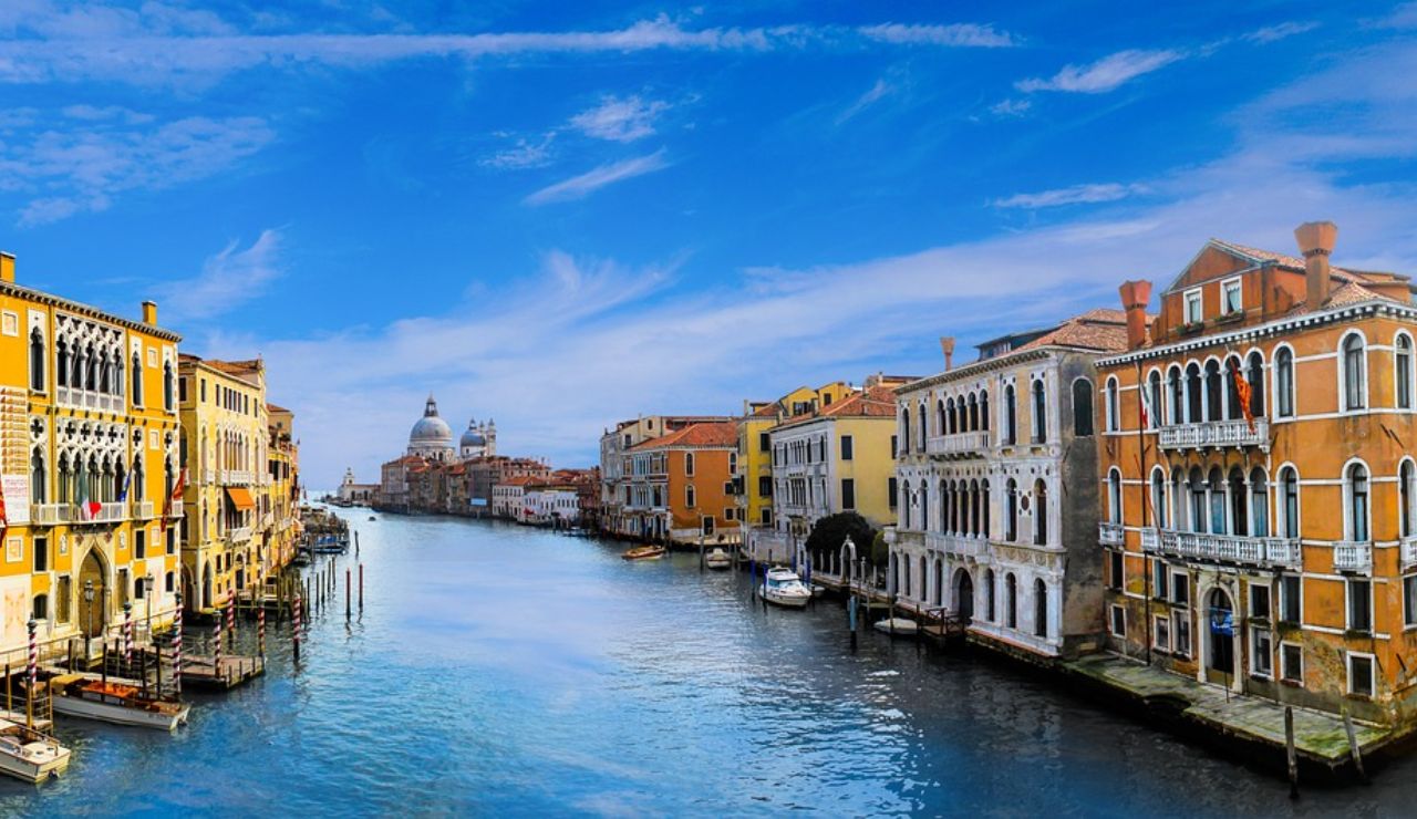 View of Venice’s Grand Canal with colorful historic buildings lining the waterway and the dome of Santa Maria della Salute in the distance under a bright blue sky.