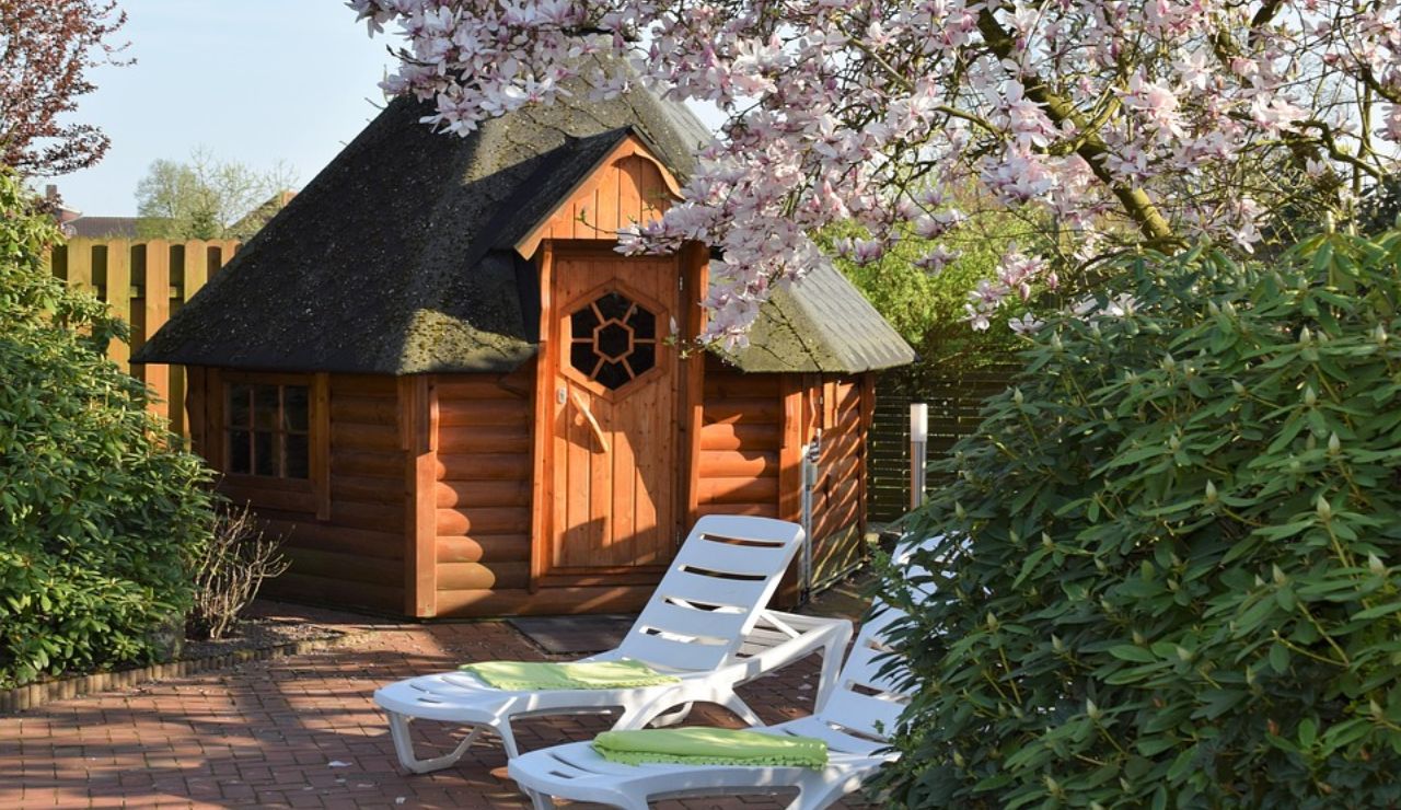 A small hexagonal wooden cabin with a shingled roof surrounded by blooming trees, bushes, and white lounge chairs on a brick patio.